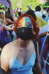 portrait of queer young woman with facemask and rainbow pride flag facepaint walking in pride parade for lgbtq rights 