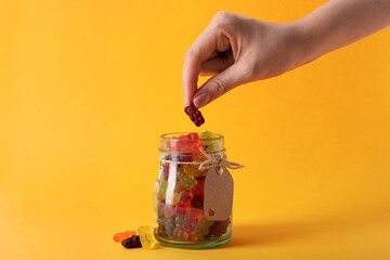 Woman taking gummy bear candy from glass jar on yellow background, closeup