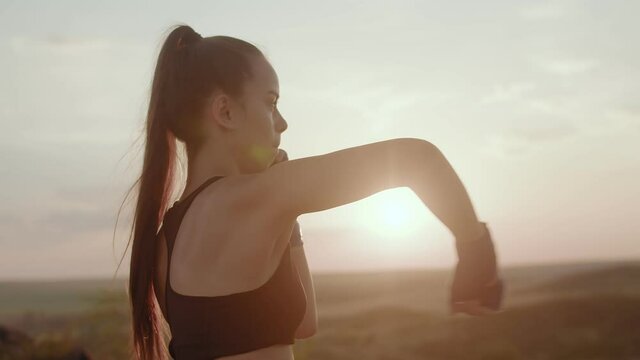Young woman on background of sunset with blue bandages on her hands kneads elbow joint, side view. Girl is warming up in nature.