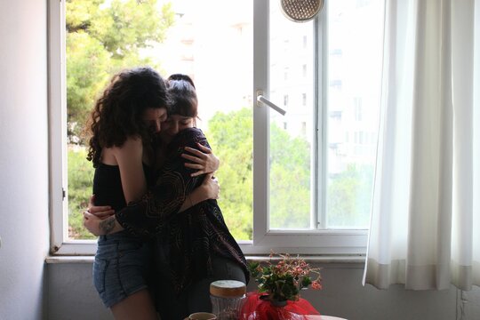 happy portrait of two women / lesbian couple in love cuddling hugging at home kitchen window *2