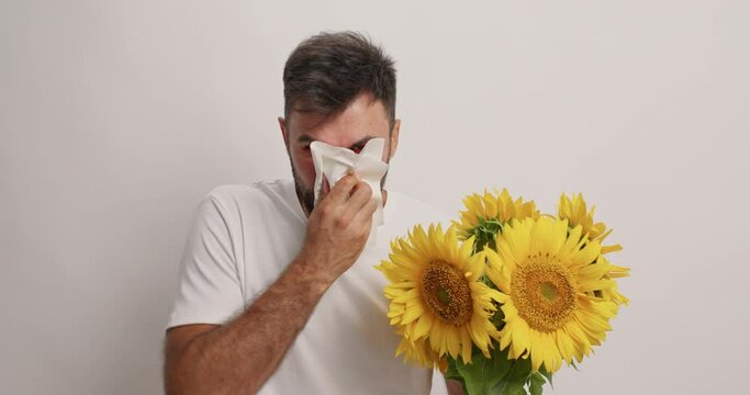 People allergy unpleasant symptoms. Displeased man reacts on allergen holds bouquet of sunflowers blows nose in tissue suffers from sneezing rhinitis has red itchy eyes needs to visit allergist