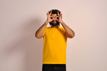 Indian man wearing yellow t-shirt doing ok gesture like binoculars eyes looking through fingers. Crazy expression.
