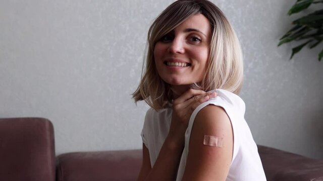 Portrait of a female smiling after getting a vaccine. Woman holding down her white shirt sleeve and showing her arm with bandage after receiving vaccination. Concept of recommended inoculation