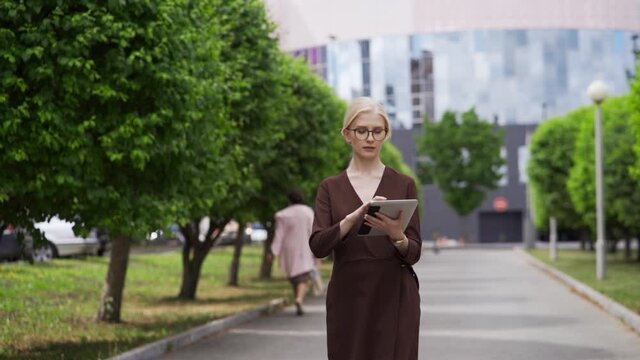 A young woman in a business suit and formal glasses walks down the street and scrolls the tablet. An important project at work. City center, capital. Slow motion, HD.