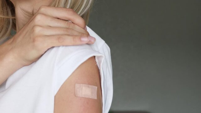 Portrait of a female smiling after getting a vaccine. Woman holding down her white shirt sleeve and showing her arm with bandage after receiving vaccination. Concept of recommended inoculation