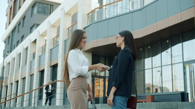 Confident young women are meeting outside office center shaking hands talking and gesturing standing outdoors in business district. People and partnership concept.