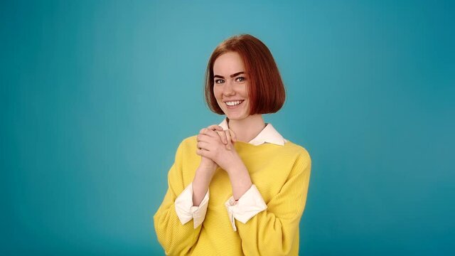 Smiling young woman with red hair and yellow pullover looks straight and joins hands back blue background slow motion close view