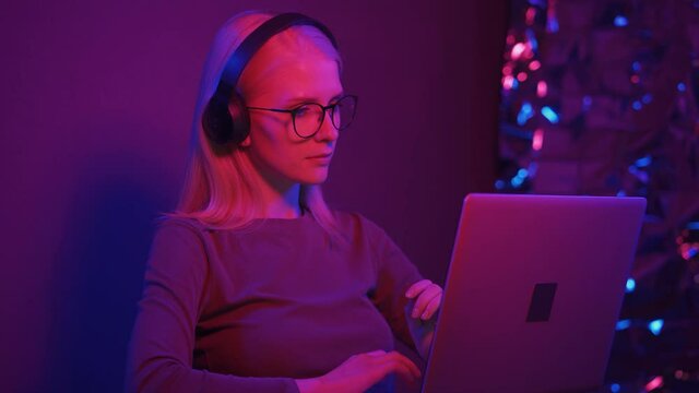 Young woman beatmaker in large headphones processes music on a laptop in neon light. Modern youth, music, heavy bass and energetic beat. Close-up, 4K UHD.