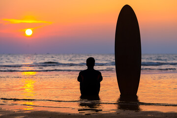 Reflection of a man watching the sunset in the sea and a surfboard on the  beach in Thailand.