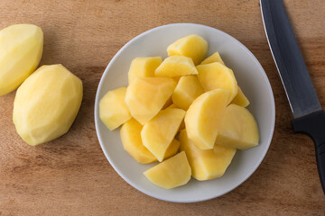 chopped raw potatoes on a white plate, with a knife and whole peeled potatoes on the background, closeup view taken from above