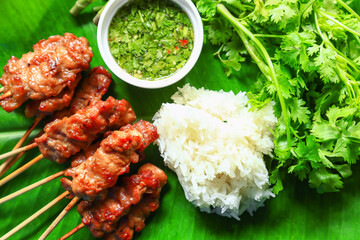grilled skewered pork and white sticky rice with coriander as a side dish with seafood sauce on a banana leaf background, local Asian Thai street food style