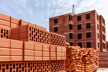 Fototapeta premium Stack of red clay brick on construction site on multistory building background.