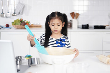 Asian little girl child is learning and enjoying for baking cookies bakery in a white bowl on wooden table in kitchen. Homemade pastry for bread. Family love and Homeschool.