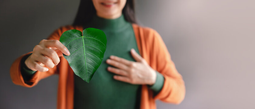 Green Energy, ESG, Renewable and Sustainable Resources. Environmental and Ecology Care Concept. Close up of Smiling Woman Holding a Heart Shape Green Leaf, presenting to Camera