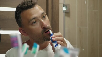Handsome guy enjoying morning oral hygiene routine alone in bathroom close-up. Happy middle-aged man brushes teeth with electric toothbrush