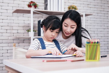 Smiling Asian mother and little girl child is drawing and Painting with wooden colored pencils on paper together in worksapce area at home. Homeschool and educational concept