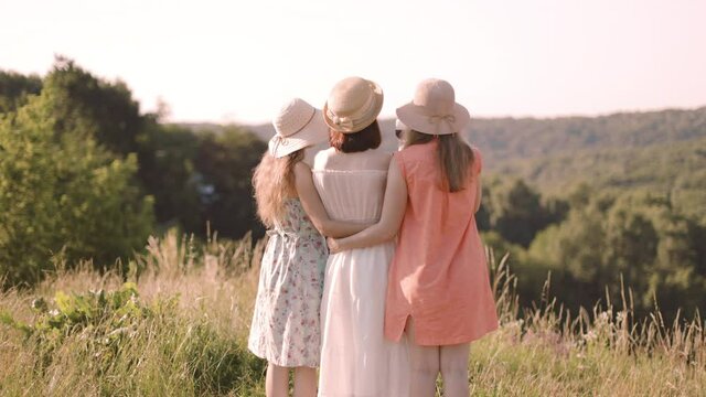Back view of company of three young female friends having fun, drink red wine, raising glasses and enjoy green hill landscape and beautiful sunset at summer picnic.