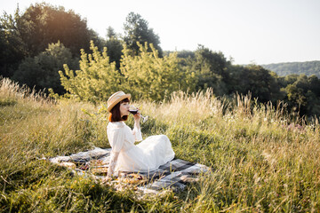 Elegant stylish pretty woman in white dress and straw hat enjoying beautiful summer sunset with red wine outdoors, sitting on the picnic blanket on top of the green hill.