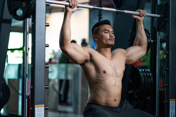 Fototapeta na wymiar Handsome shirtless adult Asian men sweating while lift up the barbell workout machine for muscle part inside of fitness gym. Bodybuilding athlete sport training for body strength and good health.