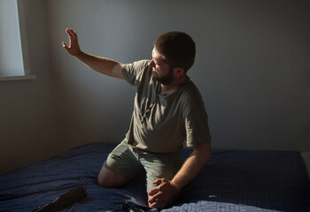 a man is sitting in a room on a bed and covers his hand from the light from the window. Next to him...