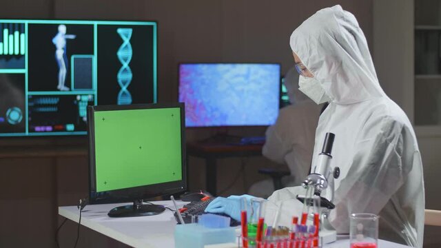 Asian Doctor In Protective Suit Working With Mock Up Green Screen Computer Display In A Modern Lab. Laboratory Assistant Workplace
