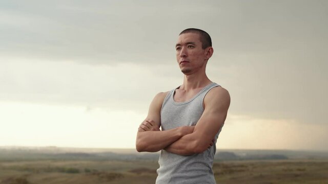 Asian man of athletic build stands in field with his arms crossed and looks ahead. Rotation shooting, closeup, side view. Concept of motivation and aspiration