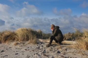 Frau in Trauer mit Händen vor dem Gesicht begraben sitzt iin einer Düne am Strand.	