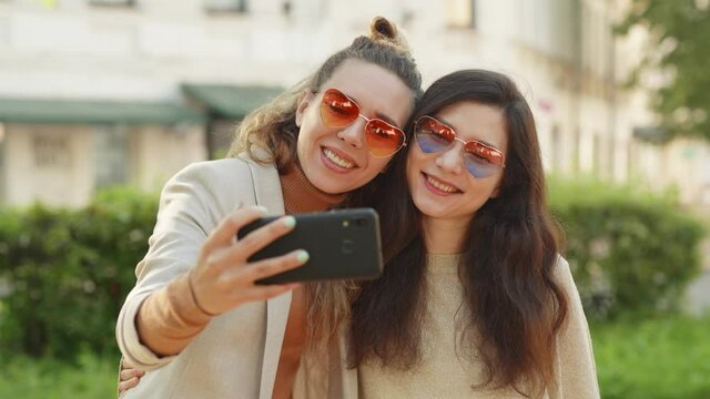 Pretty stylish girls on street background. Two young smiling hipster blond women taking selfie self portrait photos on smartphone. Positive emotions and love. LGBTQI, Pride Event, LGBT Pride Month