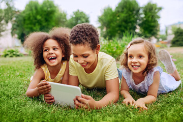 Happy little kids holding tablet PC outdoors in summer park on a green grass