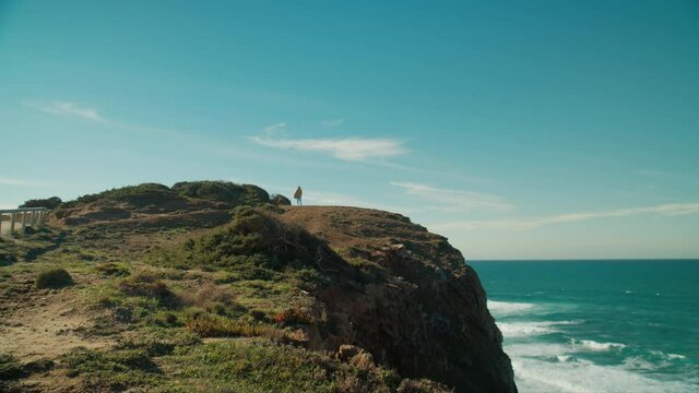 Silhouette of woman in coat flowing in strong coastal wind stand on edge of beautiful epic mountain cliff overlooking ocean. Cinematic travel mood and inspiring landscape of Portugal