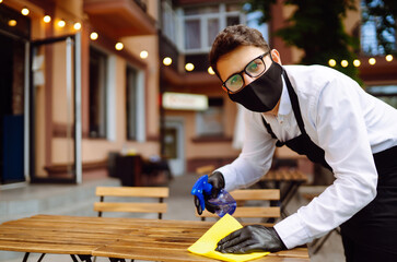 Disinfecting to prevent COVID-19. Waiter in protective face mask and gloves cleaning and disinfecting restaurant table for next customer.