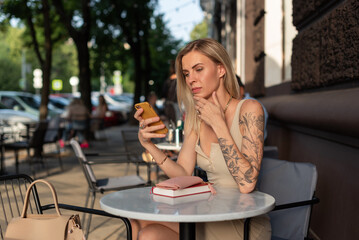 A blonde with a tattoo on her arm sits in a summer cafe and emotionally talks on the phone via video communication. Blogger concept