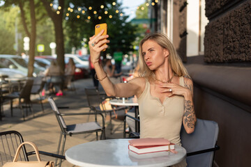 A blonde with a tattoo on her arm sits in a summer cafe and emotionally talks on the phone via video communication. Blogger concept