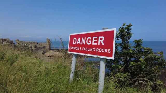 Bright red and white warning sign on the edge of a cliff ‘DANGER  EROSION AND FALLING ROCKS’.  With the ruins of St Andrews in the background.
