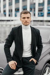 A handsome young man in a black suit is standing near a sports car. A man with a watch on his hands in a dark business suit