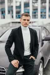 A handsome young man in a black suit is standing near a sports car. A man with a watch on his hands in a dark business suit