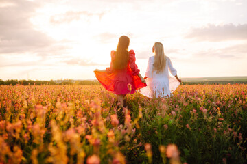 Portrait of two fair-haired girls in fashionable and stylish dresses in the blooming field. Nature, vacation, relax and lifestyle. Fashion concept.
