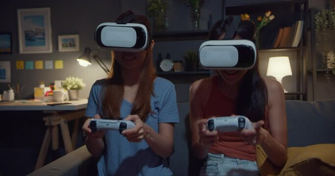 Attractive Asia couple ladies enjoy happy moment with vr headset or virtual reality glasses and controller gamepad experience playing video game site on couch living room in home at dark night.
