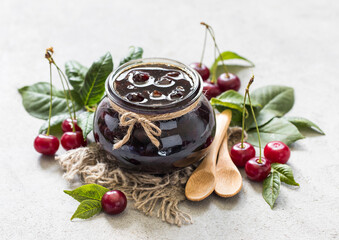 Cherry jam in a glass jar on a linen napkin with wooden spoons on a light background in rustic style