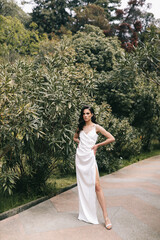 Beautiful elegant young woman brunette bride in a wedding dress posing walking alone one among the architectural columns and green plants in the garden of the city park outdoors, selective focus