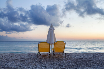 Holiday time, vacation, Panorama of the pebble coast with white pebbles with two yellow deck chairs and calm azure blue sea at dusk