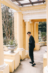 A beautiful fashionable stylish cool young man in an elegant wedding suit poses and walks alone among plants and palm trees in an old park in nature outdoors, selective focus