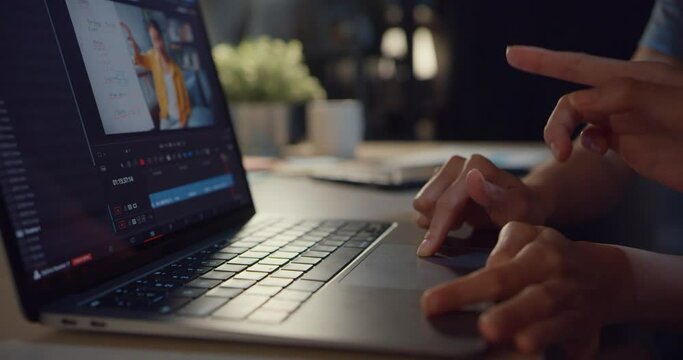 Young Asia ladies blogger watching footage on laptop sharing ideas with friend and working on video editing on computer doing montage and color correction in living room from home at night.