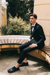 A beautiful fashionable stylish cool young man in an elegant wedding suit poses and walks alone among plants and palm trees in an old park in nature outdoors, selective focus