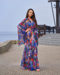 Elegant caucasian woman with wavy brunette hair in vivid blue colorful dress walking outdoors on a sunny summer day at pavement against view on s sea water