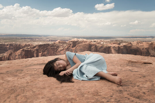 Asian girl in blue dress laying down and dreaming on edge of crater on red soail in Canyonlands National Park, USA