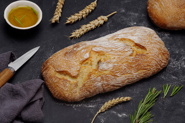 Crispy artisan ciabatta bread, olives, olive oil, rosemary, wheat branch, knife on black background.  - obrazy, fototapety, plakaty