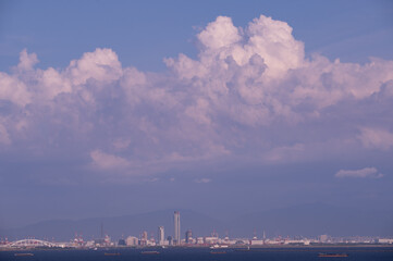 Fototapeta premium 夏の夕暮れ、大阪湾に入道雲が浮かぶ。夕陽がさして雲が少しオレンジ色に染まる。海の向こう岸、遠くには大阪の南港と高層ビル群が見える。神戸からの対岸の眺め。