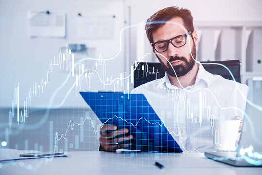 Handsome beard businessman trader in glasses is sitting at workplace and working with documents to continue trading operations. Forex charts. Concept of analyzing stock market behavior