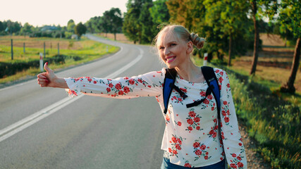 Traveler woman hitchhiking on a sunny road and walking. Young happy backpacker woman looking for a ride to start a journey on a sunlit country road.